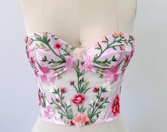 May include: A pink strapless corset top with floral embroidery. The corset features a sweetheart neckline and is adorned with embroidered flowers in shades of pink, red, and green. The garment is displayed on a mannequin.