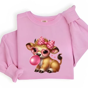 Puede incluir: Sudadera rosa con un diseño de vaca de dibujos animados. La vaca tiene un lazo rosa con lunares blancos, gafas en forma de corazón y una burbuja de chicle. La vaca es marrón con cuernos pequeños. También se incluyen pequeños corazones.