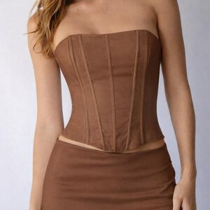 May include: A brown strapless corset top and matching mini skirt. The corset features vertical seams for structure. The outfit is a solid, warm brown color, creating a cohesive and stylish ensemble. The skirt sits at the natural waist.