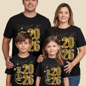 Puede incluir: Cuatro camisetas negras con un diseño de purpurina dorada. El diseño presenta los números "2026" y las palabras "Happy New Year" en una fuente cursiva, junto con estrellas y líneas doradas. Las camisetas son usadas por una familia de cuatro.
