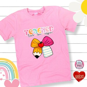 May include: Pink t-shirt with the word "TEACHER" in colorful letters. Below the word is "teach love inspire". The shirt features a pencil and a pink bow with an apple in the center.