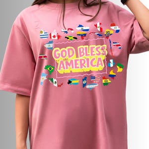 Puede incluir: Camiseta rosa claro con el texto "GOD BLESS AMERICA" en amarillo y rosa. La camiseta presenta un diseño de banderas y contornos de países alrededor del texto, incluida la bandera estadounidense.