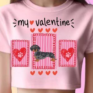 Puede incluir: Top corto rosa con temática de San Valentín. La camiseta presenta el texto "my valentine" sobre una ilustración de perro salchicha y tres diseños en forma de corazón con huellas de patas. El diseño es en rojo y negro.