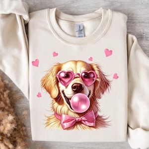 Puede incluir: Sudadera color crema con un golden retriever con gafas de sol en forma de corazón y una pajarita rosa. El perro está soplando una burbuja rosa y está rodeado de corazones rosas. Un diseño divertido y caprichoso.