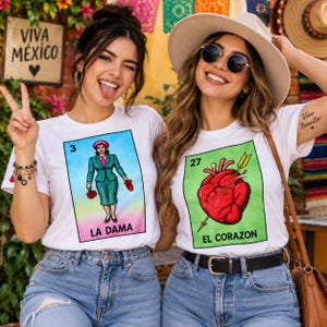 May include: Two white t-shirts featuring colorful Loteria card graphics. The left shirt displays a woman in a teal suit labeled LA DAMA with the number 3. The right shirt shows a red heart pierced by an arrow labeled EL CORAZON with the number 27.