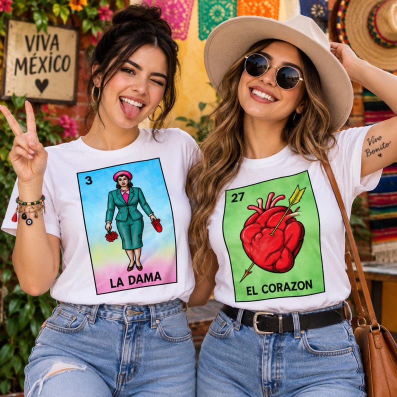 May include: Two white t-shirts featuring colorful Loteria card graphics. The left shirt displays a woman in a teal suit labeled LA DAMA with the number 3. The right shirt shows a red heart pierced by an arrow labeled EL CORAZON with the number 27.