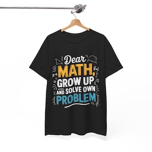 Könnte beinhalten: Schwarzes T-Shirt mit dem Text "Dear Math, Grow Up, and Solve Own Problem" in Weiß, Gelb und Blau. Das T-Shirt hängt an einem Holzbügel.