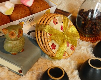 May include: An assortment of items including baked goods, a decorative owl figurine, and small black bowls. A woven basket with a gold ribbon and a glass jar filled with dark granules are also present. The scene is set on a soft, textured surface.