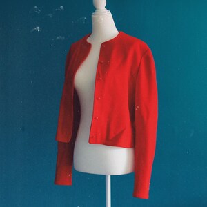 May include: A bright red, button-down jacket with long sleeves. The jacket is displayed on a white mannequin against a teal background. The jacket has a round neckline and is made of a smooth fabric.