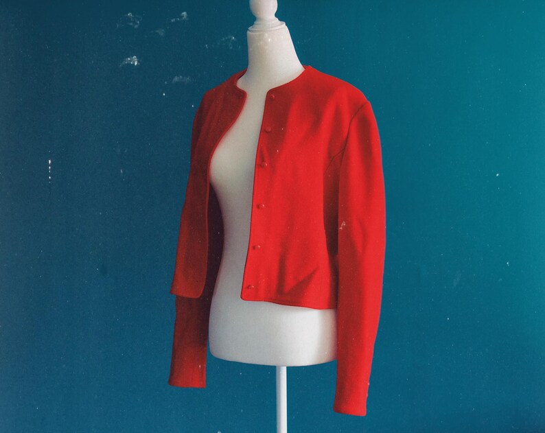 May include: A bright red, button-down jacket with long sleeves. The jacket is displayed on a white mannequin against a teal background. The jacket has a round neckline and is made of a smooth fabric.