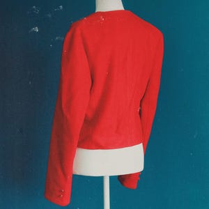 May include: A bright red blazer with long sleeves and button details on the cuffs. The jacket is displayed on a white mannequin torso against a teal background. The blazer's back is visible, showcasing its tailored design.