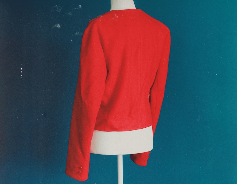May include: A bright red blazer with long sleeves and button details on the cuffs. The jacket is displayed on a white mannequin torso against a teal background. The blazer's back is visible, showcasing its tailored design.