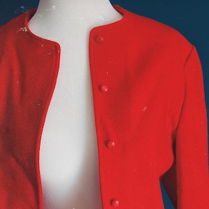 May include: A bright red jacket with a round collar and three matching buttons. The jacket is displayed on a white mannequin against a dark blue background. The jacket's fabric appears to be a smooth, possibly wool, material.