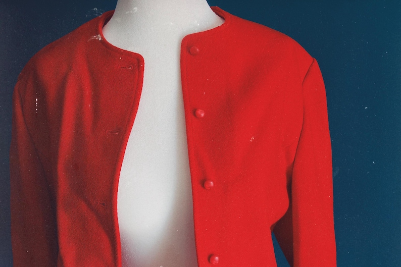 May include: A bright red jacket with a round collar and three matching buttons. The jacket is displayed on a white mannequin against a dark blue background. The jacket's fabric appears to be a smooth, possibly wool, material.