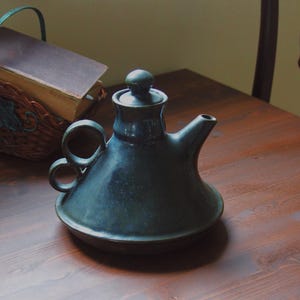 May include: A dark teal ceramic teapot with a rounded base, a spout, and a small handle on the lid. The teapot has two loop handles and sits on a wooden table. A basket with a book is in the background.