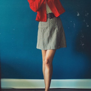 May include: A person wearing a red blazer, a black belt, and a plaid skirt. The person is also wearing red flats. The background is a solid blue wall.