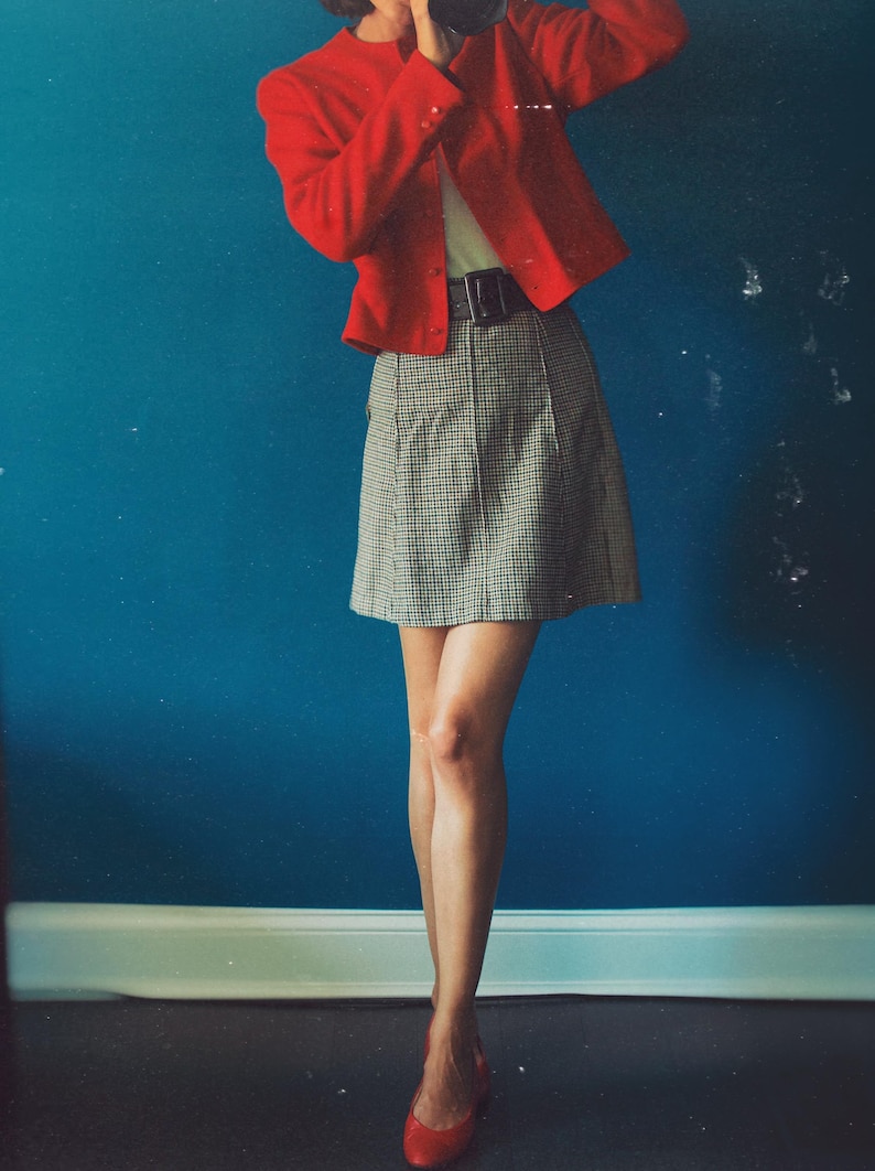 May include: A person wearing a red blazer, a black belt, and a plaid skirt. The person is also wearing red flats. The background is a solid blue wall.