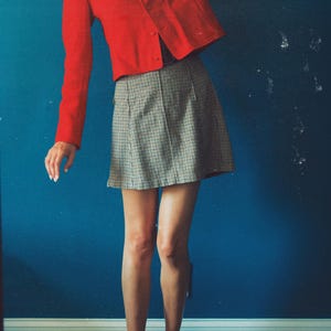 May include: A red, long-sleeved jacket with a round neckline and a matching skirt in a black and white checkered pattern. The outfit is paired with red flats. The background is a solid blue wall.
