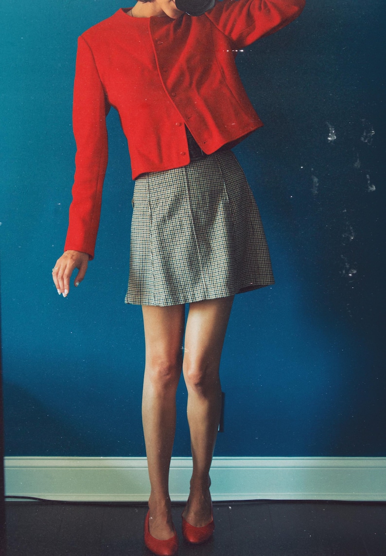 May include: A red, long-sleeved jacket with a round neckline and a matching skirt in a black and white checkered pattern. The outfit is paired with red flats. The background is a solid blue wall.