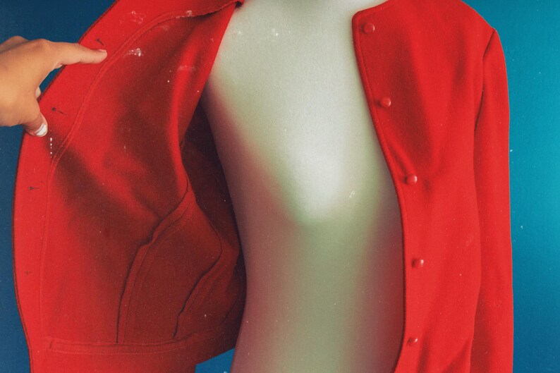 May include: A bright red jacket with a button-up front and two pockets. The jacket is displayed on a mannequin against a blue background. The inside of the jacket is visible, showing the lining and construction.