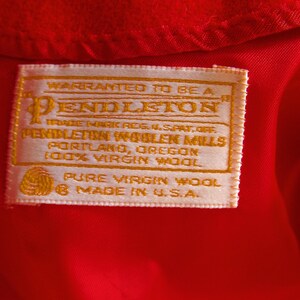 May include: Close-up of a red fabric with a rectangular label. The label is cream-colored with gold text, reading "Pendleton" and "100% Virgin Wool." It also states "Made in U.S.A."