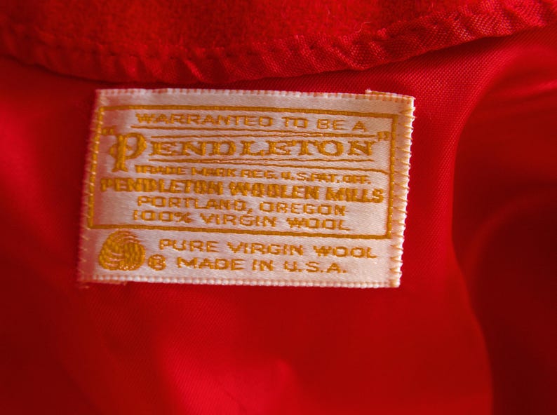 May include: Close-up of a red fabric with a rectangular label. The label is cream-colored with gold text, reading "Pendleton" and "100% Virgin Wool." It also states "Made in U.S.A."