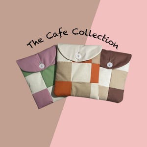 May include: Three quilted fabric cases in various colours. The cases are in shades of pink, green, beige, orange, and brown. Each case has a flap closure with a white or brown button. The text "The Cafe Collection" is above the cases.