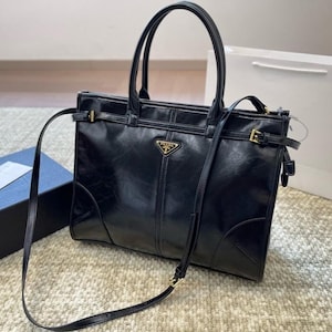 Puede incluir: Bolso tote de cuero negro con asas superiores y bandolera desmontable. El bolso presenta herrajes dorados, incluyendo hebillas y un emblema con el logotipo. El bolso tiene forma rectangular y un diseño estructurado.