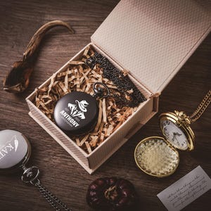 May include: A collection of pocket watches, one gold and two silver, and a black watch in a gift box. The black watch has the name "ANTHONY" and a logo. The gold watch is open, revealing the clock face.