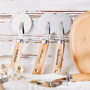 Könnte beinhalten: Drei Pizzaschneider mit Holzgriffen, graviert mit dem Namen "Anderson" und Edelstahlklingen. Ein Holzschneidebrett und ein Pizzaschneider sind ebenfalls sichtbar. Die Artikel sind auf einer weißen Oberfläche angeordnet.