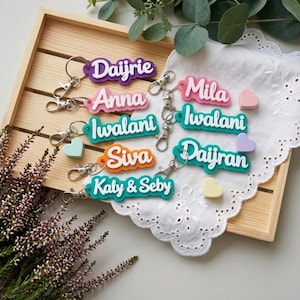 May include: A collection of colorful personalized name keychains with silver hardware. The keychains feature names in various colors, including purple, pink, teal, orange, and teal. Small heart-shaped charms in pastel colors are also present.