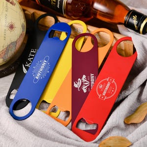 Könnte beinhalten: Eine Sammlung farbenfroher Flaschenöffner in Schwarz, Blau, Gelb, Orange und Rot. Jeder Öffner hat ein einzigartiges Design mit dem Namen "ANTHONY" und anderem Text. Eine Whiskyflasche und ein Globus befinden sich im Hintergrund.