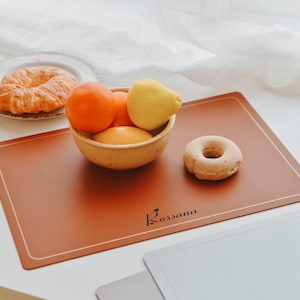 Può includere: Un sottopiatto rettangolare, color ruggine, con un sottile bordo color crema. Una piccola ciotola piena di arance e un limone si trova sul tappetino, insieme a una ciambella. La parola "Rossana" è stampata in nero nell'angolo in basso a destra.