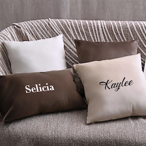 May include: Four decorative cushions on a sofa. Two rectangular cushions are brown, with names printed on them. Two square cushions are white and beige. A book with the title "Raison de Soleil" is visible.