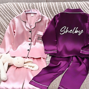 May include: Two sets of personalized satin pajamas. One set is pink with black trim and the name "Tang" embroidered on the pocket. The other set is purple with white trim and the name "Shelby" on the back. A white stuffed bunny is in the foreground.