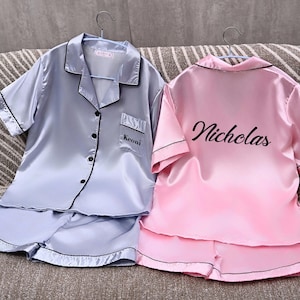 Pode incluir: Dois conjuntos de pijamas de cetim, um azul claro e outro rosa, cada um com acabamento preto. O conjunto azul claro tem "Keoni" bordado no bolso. O conjunto rosa tem "Nicholas" impresso nas costas. Ambos os conjuntos incluem uma camisa com botões e calções.