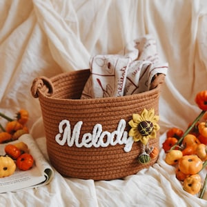 May include: A brown woven basket with the name "Melody" in white script. A yellow sunflower and small pumpkins decorate the basket. The basket contains a folded cloth and is surrounded by small orange pumpkins.