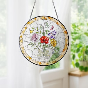 Puede incluir: Vidriera redonda que representa un ramo floral con una amapola roja, un iris morado y flores amarillas. El dise&ntilde;o incluye mariposas y el texto "Nana's Garden". Tiene un borde negro y una cadena para colgar.