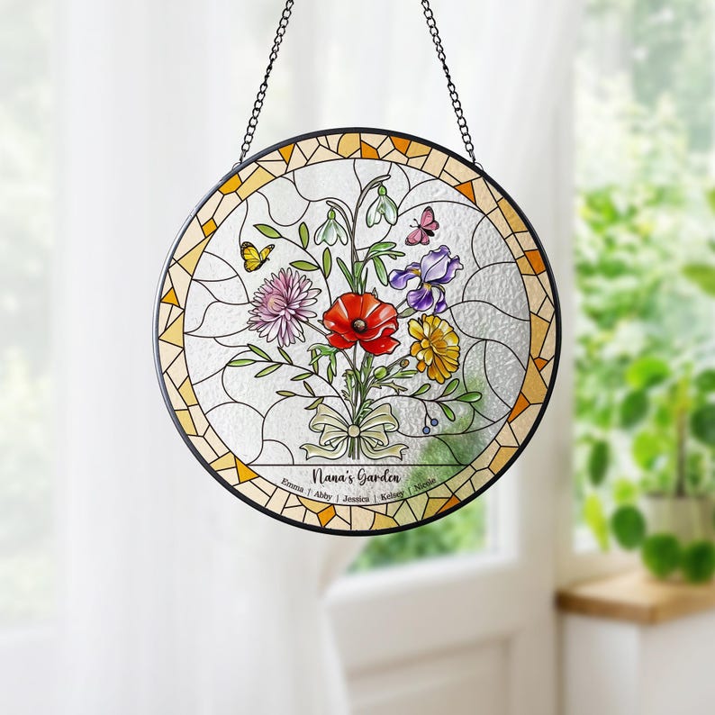 Puede incluir: Vidriera redonda que representa un ramo floral con una amapola roja, un iris morado y flores amarillas. El dise&ntilde;o incluye mariposas y el texto "Nana's Garden". Tiene un borde negro y una cadena para colgar.