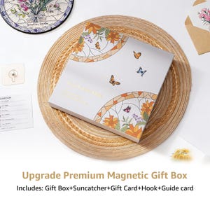 Puede incluir: Una caja de regalo blanca con un dise&ntilde;o floral y de mariposas, sobre una estera tejida. La caja presenta el texto "Love Grows Here" en letras doradas. La imagen tambi&eacute;n incluye un atrapasoles, una tarjeta de regalo, un gancho y una gu&iacute;a.