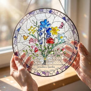 Puede incluir: Una vidriera redonda con un dise&ntilde;o de ramo floral. El ramo presenta flores azules, amarillas, rojas y rosas, con mariposas. El texto "Grandma's Garden" se muestra en la parte inferior, junto con nombres. Una cadena est&aacute; adjunta para colgar.