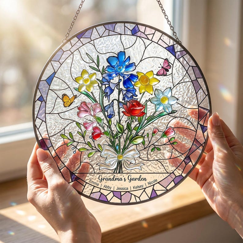 Puede incluir: Una vidriera redonda con un dise&ntilde;o de ramo floral. El ramo presenta flores azules, amarillas, rojas y rosas, con mariposas. El texto "Grandma's Garden" se muestra en la parte inferior, junto con nombres. Una cadena est&aacute; adjunta para colgar.