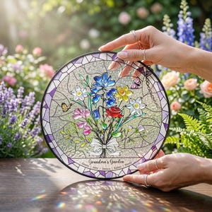 Puede incluir: Vidriera redonda con un ramo de flores con flores azules, amarillas, rosas y rojas. Se muestra el texto "Grandma's Garden", junto con nombres. La ventana tiene un borde morado y est&aacute; sostenida por manos.