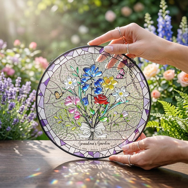 Puede incluir: Vidriera redonda con un ramo de flores con flores azules, amarillas, rosas y rojas. Se muestra el texto "Grandma's Garden", junto con nombres. La ventana tiene un borde morado y est&aacute; sostenida por manos.