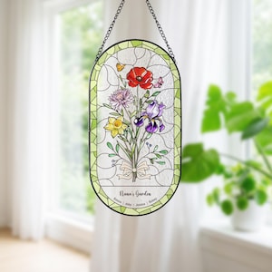 Puede incluir: Vidriera con un ramo de flores coloridas, incluyendo una amapola roja, un iris morado y un narciso amarillo. El panel ovalado est&aacute; enmarcado en negro y cuelga de una cadena. El texto "Nana's Garden" se muestra en la parte inferior.
