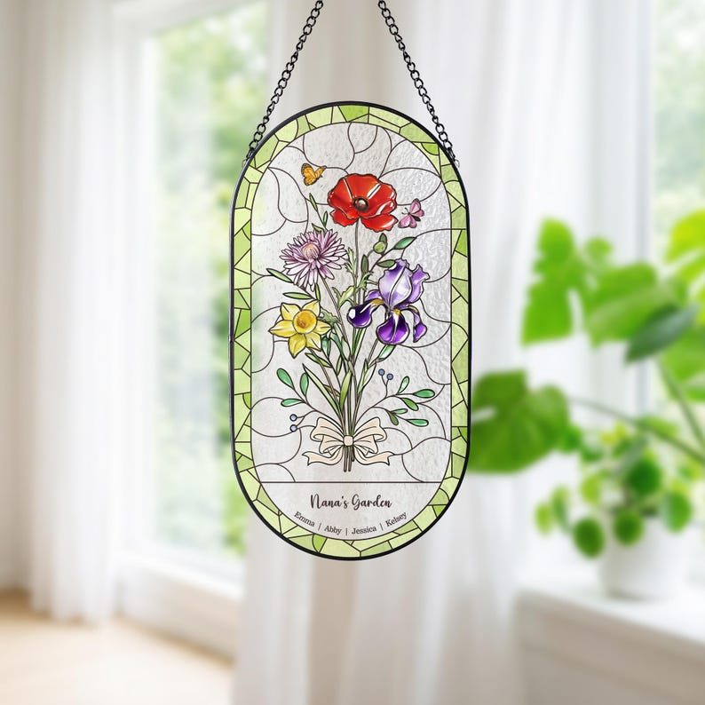 Puede incluir: Vidriera con un ramo de flores coloridas, incluyendo una amapola roja, un iris morado y un narciso amarillo. El panel ovalado est&aacute; enmarcado en negro y cuelga de una cadena. El texto "Nana's Garden" se muestra en la parte inferior.