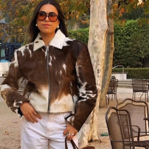 Könnte beinhalten: Eine braun-weiße Kuhfelljacke mit weißem Kragen und silbernem Reißverschluss. Die Jacke hat ein klassisches Design und wird mit einer weißen Hose kombiniert. Die Person trägt orange getönte Sonnenbrillen.