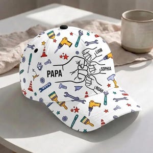 Può includere: Un cappellino da baseball bianco con un motivo ripetuto di attrezzi da costruzione colorati come trapani, chiavi inglesi e pennelli. La parte anteriore mostra un grafico di un pugno contro pugno con il testo PAPA, LIAM, SOPHIA e NOAH.