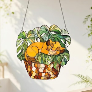 May include: Stained glass art piece featuring a sleeping lion cub nestled in a bed of green leaves. The cub is a warm yellow-orange color, and the leaves are a mix of green and white. The planter is a mosaic of brown, orange, and yellow pieces.