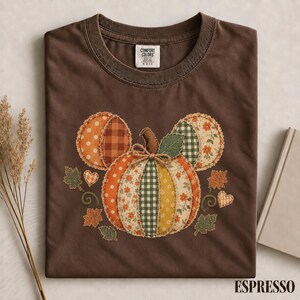 May include: A dark brown short-sleeve t-shirt featuring a central graphic of a pumpkin constructed from various patterned fabric patches in orange, yellow, and green. The pumpkin has a twine bow and is surrounded by small hearts and leaves.